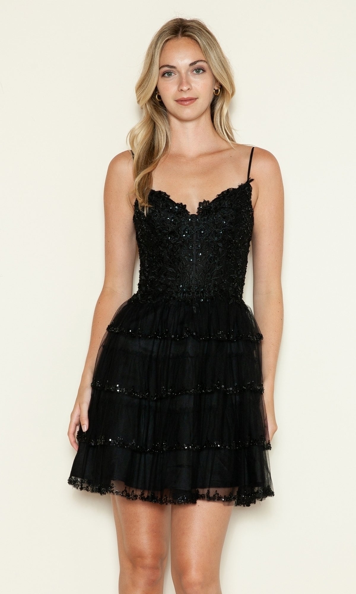 A woman stands against a plain light background, wearing the Short Sparkly A-Line Homecoming Dress 9592—black, sleeveless, with sequin details, thin straps, and a tiered mini skirt.
